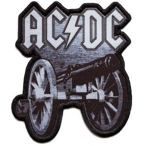 以下が含まれることがあります： AC/DCのバンド名と稲妻のグラフィックが特徴の白黒刺繍パッチ。テキストの下には大砲の詳細なイラストがあります。パッチは黒い縁取りがされています。