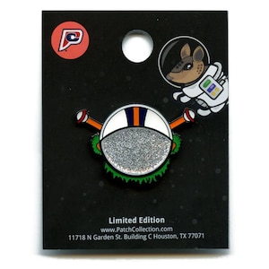 May include: A limited edition pin badge featuring a cartoon armadillo astronaut in a spacesuit. The badge has a white and silver glitter face, orange arms, and green accents. The badge is on a black backing card with a logo.