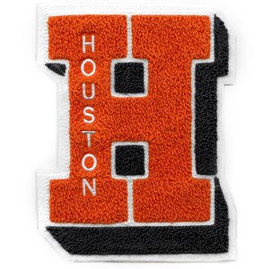 Könnte beinhalten: Orangefarbener Filzbuchstabe H mit schwarzem Umriss und weißen Nähten. Der Buchstabe ist von einem weißen Rand umgeben. Das Wort "HOUSTON" ist vertikal auf der linken Seite des Buchstabens gestickt.