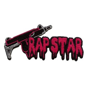 Könnte beinhalten: Ein gestickter Aufnäher mit einer schwarzen und silbernen Maschinenpistole und dem Wort "RAPSTAR" in tropfenden pinkfarbenen Buchstaben.