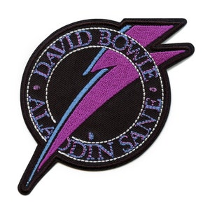 May include: Embroidered patch with a black circular design and a purple lightning bolt. The patch features the words "DAVID BOWIE" and "ALADDIN SANE" in white stitching. The lightning bolt is outlined in light blue.