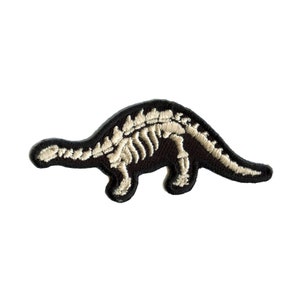 Peut inclure: Un patch brodé noir représentant un squelette de dinosaure. Le dinosaure est tourné vers la droite et a un long cou et une longue queue.
