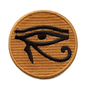 May include: A round, gold-colored patch with a black embroidered image of the Eye of Horus, an ancient Egyptian symbol.