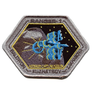 Puede incluir: Un parche bordado hexagonal con un borde plateado. El parche presenta un fondo marrón oscuro con una nave espacial azul y amarilla en el centro. La nave espacial está volando junto a una luna marrón y gris. El texto "RANGER 1" está en la parte superior del parche, y "ASTEROID CAPTURE VEHICLE" está en la parte inferior.