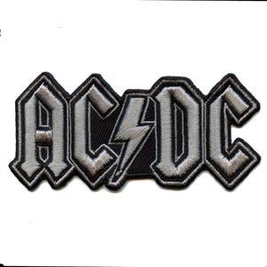 May include: A black and silver embroidered patch featuring the word "AC/DC" in a bold, stylised font. The lightning bolt is a prominent feature in the design.
