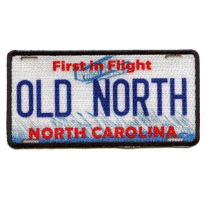Może przedstawiać: Naszywka haftowana w kolorze białym i niebieskim z tekstem "Old North" i "North Carolina" w kolorze niebieskim. Tekst "First in Flight" jest w kolorze czerwonym nad głównym tekstem. Naszywka jest otoczona czarną ramką.