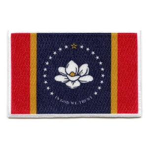 Op de afbeelding: Een geborduurde patch in blauw en rood met een witte magnolia bloem in het midden. De patch heeft een cirkel van witte sterren rond de bloem en de woorden "In God We Trust" onderaan.
