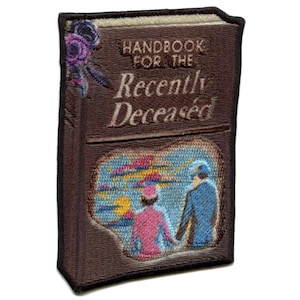 May include: A brown book with the title "Handbook for the Recently Deceased" in gold lettering. The cover features an illustration of a couple walking away from a city skyline.