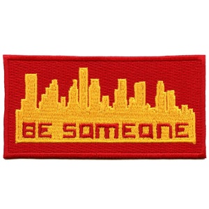 Könnte beinhalten: Roter gestickter Aufnäher mit einer gelben Skyline und dem Text "BE SOMEONE" in Gelb.
