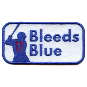 以下が含まれることがあります： 野球選手のシルエットと「Bleeds Blue」というテキストが刺繍された青と白のワッペン。選手のユニフォームの番号は赤で17です。