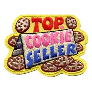 May include: Yellow embroidered patch with the text "TOP COOKIE SELLER" in colorful letters. The patch features a cartoon image of a cookie stand with cookies around it.