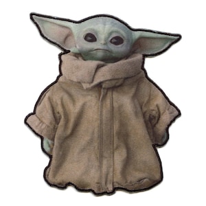 Op de afbeelding: Een bruine en groene pluche patch van een personage uit de Star Wars-franchise, bekend als "The Child" of "Baby Yoda". Het personage draagt een bruine robe en heeft grote oren.