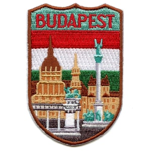 May include: Embroidered patch in the shape of a brown shield. The shield features a cityscape with buildings and a column, with the Hungarian flag behind it. The text "BUDAPEST" is embroidered in red at the top of the shield.