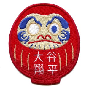 Può includere: Bambola daruma rossa e bianca ricamata con occhi blu e il testo "大谷翔平" in giapponese.
