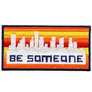 May include: An embroidered patch featuring a white cityscape silhouette against a backdrop of orange, yellow and red stripes. The text "BE SOMEONE" is embroidered in white below the cityscape.