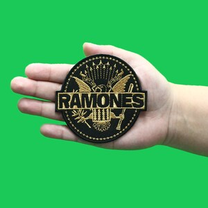 May include: Black and gold embroidered patch with the word "RAMONES" in a banner across the center. The patch features a stylized eagle with wings spread and a crown on its head. The eagle is surrounded by a circle of stars.