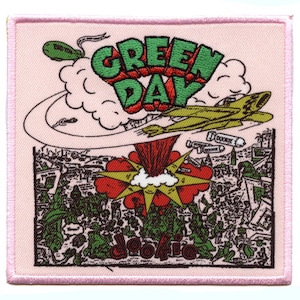 Puede incluir: Un parche de tela rosa con una imagen bordada de un avión de dibujos animados que lanza bombas sobre una ciudad. El texto "GREEN DAY" está en letras verdes grandes sobre la imagen. El texto "Dookie" está en letras negras debajo de la imagen.