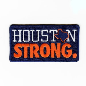 May include: A blue embroidered patch with the text "HOUSTON STRONG" in white and orange lettering. A small outline of the state of Texas is in the middle of the text "HOUSTON".