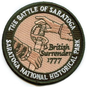 Puede incluir: Un parche bordado verde y marrón con el texto "The Battle of Saratoga, Saratoga National Historical Park" y una representación de un soldado británico entregando su espada a un soldado estadounidense. El texto "British Surrender 1777" también está incluido.