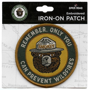 May include: A round, embroidered iron-on patch featuring Smokey Bear. The patch is yellow with a green border and the text "Remember, Only You Can Prevent Wildfires". Smokey Bear is wearing his iconic ranger hat with the word "Smokey" on it.