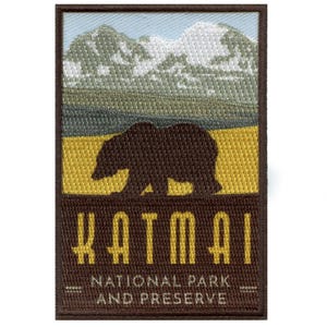 May include: Embroidered patch featuring a brown bear silhouette against a backdrop of mountains and a yellow and green landscape. The text "Katmai National Park and Preserve" is displayed in yellow.