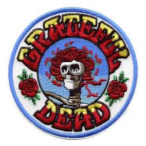 May include: A circular embroidered patch with a white background and a blue border. The patch features a red, yellow, and black stylized text that reads "Grateful Dead". In the center of the patch is a cartoon image of a skull with a red rose crown, a blue background, and a brown branch.