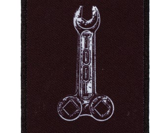 Könnte beinhalten: Ein schwarzer Stoff-Patch mit einem weißen Schraubenschlüssel-Design. Der Schraubenschlüssel trägt die Aufschrift "TOOL".