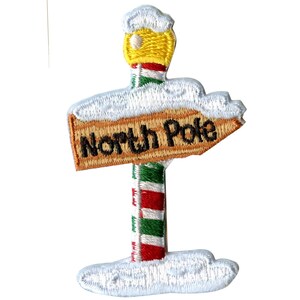 Pode incluir: Um remendo bordado que apresenta uma placa de madeira com as palavras "Polo Norte" apontando para a direita. A placa é coroada com um poste de pirulito com uma estrela amarela no topo. A placa é cercada por neve branca.