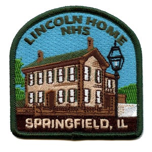 Op de afbeelding: Een geborduurde patch met een groene rand, die een huis met twee verdiepingen met een bruin dak en witte ramen toont. Het huis staat tegen een blauwe lucht met groene bomen aan beide kanten. De tekst "LINCOLN HOME NHS SPRINGFIELD, IL" is in bruin en wit geborduurd op de patch.