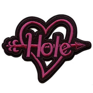 Puede incluir: Parche bordado en negro y rosa con una flecha que atraviesa un corazón y la palabra "Hole" dentro del corazón.