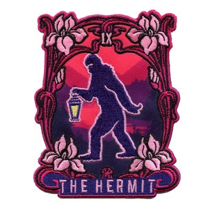 May include: An embroidered patch featuring a silhouette of a Bigfoot figure holding a lantern, walking through a pink and purple mountain landscape. The patch is framed with pink flowers and the text "THE HERMIT" is at the bottom.