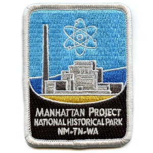 May include: A white and gray embroidered patch with a blue background. The patch features a stylized image of a nuclear reactor with a stylized atom symbol above it. The text "MANHATTAN PROJECT NATIONAL HISTORICAL PARK NM-TN-WA" is printed in black on a brown background.