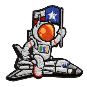 May include: An embroidered patch featuring a cartoon astronaut holding a Texas flag, sitting on a space shuttle. The astronaut is wearing a white spacesuit with an orange helmet and a colorful patch on the chest. The space shuttle is gray and black.