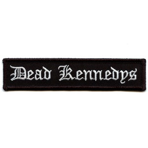 May include: A rectangular, dark brown patch with the words "Dead Kennedys" embroidered in white, gothic-style lettering. The patch has a black border and is likely designed to be sewn onto clothing or accessories.