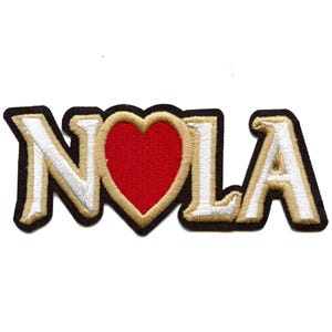 Puede incluir: Parche bordado con el texto "NOLA" en letras doradas y blancas con un corazón rojo en el centro.
