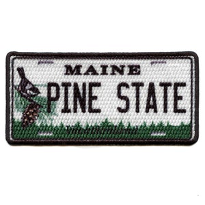 Pode incluir: Um remendo bordado em preto e branco com fundo verde. O remendo apresenta um pinheiro estilizado com um pássaro empoleirado em um galho. O texto "MAINE PINE STATE" é exibido no remendo.