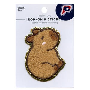 May include: A brown plush iron-on patch with a gold glitter outline. The patch features a cartoon-style brown bear with a pink nose. The text "DESIGN Loft IRON-ON & STICKER" is printed on the packaging.