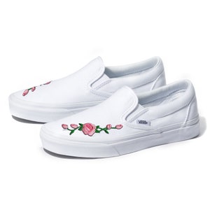 May include: White canvas slip-on Vans shoes with pink rose embroidery. The shoes have a white rubber sole and the Vans logo is visible on the side.