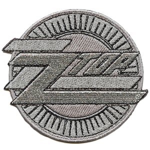 Pode incluir: Um remendo bordado prateado com a palavra "ZZ Top" em uma fonte estilizada, cercado por um design circular de linhas radiantes.
