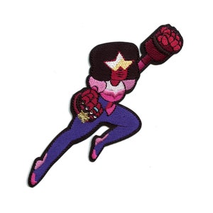 Op de afbeelding: Een geborduurde patch met een cartoonfiguur in een roze en paarse outfit, met een rode edelsteen op hun voorhoofd en een ster op hun borst. Het personage houdt een rood wapen vast en staat in een springende pose.