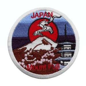May include: Embroidered patch featuring a stylized image of Mount Fuji in Japan. The patch is white with a red rising sun, a brown mountain, and a blue sky. The text "JAPAN" and "Mount Fuji" are embroidered in red.