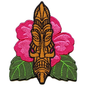 Pode incluir: Uma máscara tiki marrom com um design esculpido é cercada por flores de hibisco rosa e folhas verdes. A máscara tem um estilo polinésio tradicional.
