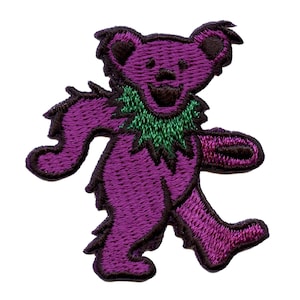 Puede incluir: Parche bordado morado de un oso bailando que lleva una guirnalda verde. El oso tiene la nariz y los ojos marrones.