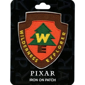 May include: A brown, orange, green, and yellow embroidered iron-on patch with a white border. The patch features a stylized pine tree with a yellow "W" inside and the text "WILDERNESS EXPLORER" around the tree. The text "PIXAR IRON ON PATCH" is printed below the patch.