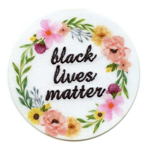 May include: A white circular patch with a floral wreath border. The wreath is made of pink, yellow, and green flowers. The text "black lives matter" is embroidered in black in the center of the patch.