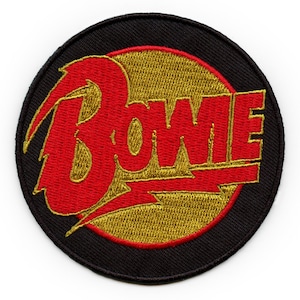 Puede incluir: Parche bordado circular con el nombre "Bowie" en rojo, con un rayo, sobre un círculo dorado y fondo negro. Ideal para ropa o accesorios.