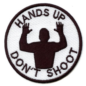 Peut inclure: Écusson brodé blanc avec une silhouette noire d'une personne les mains levées en l'air. Le texte "Hands Up Don't Shoot" est écrit autour de la silhouette.