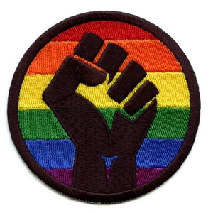 Pode incluir: Um remendo circular bordado com um punho preto em relevo no centro, rodeado por um padrão de listras arco-íris. As cores do arco-íris são vermelho, laranja, amarelo, verde, azul, anil e violeta.