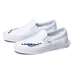 May include: White canvas slip-on Vans shoes with blue floral embroidery on the side. The shoes have a black Vans logo on the side.