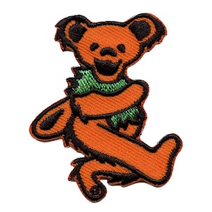 May include: Orange and black embroidered patch of a cartoon bear wearing a green scarf. The bear is standing on its hind legs with its arms crossed and its head tilted to the side.